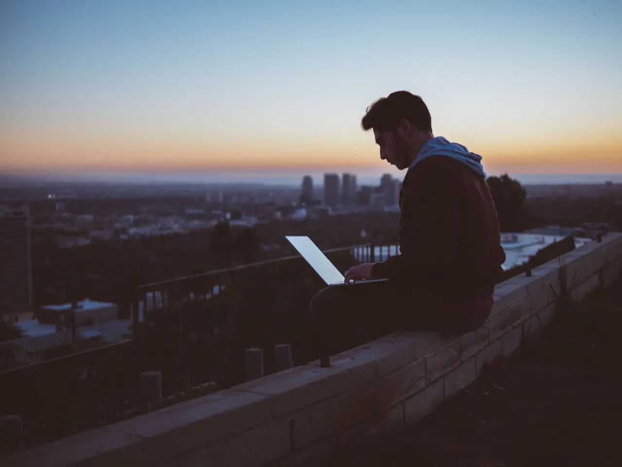 A person working on a laptop on a rooftop ledge as the city lights come on at dusk